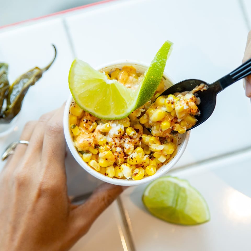 Hands holding a cup of Mexican street corn (Esquites) with lime wedges and a spoon.