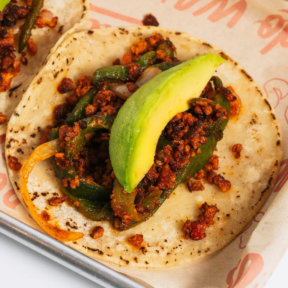 A Chido Taco with chorizo, green peppers, onions, and a slice of avocado on a corn tortilla.