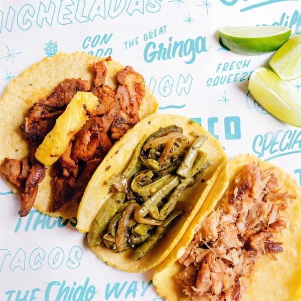 Three Chido Taco tacos: al pastor with pineapple, rajas, and carnitas, with lime wedges.