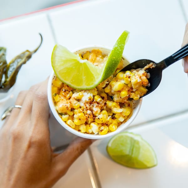 Hands holding a cup of Mexican street corn (Esquites) with lime wedges and a spoon.