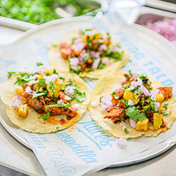 Three Chido Taco al pastor tacos with pineapple, onions, and cilantro on a metal tray.