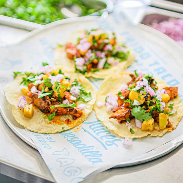 Three Chido Taco al pastor tacos with pineapple, red onion, and cilantro on a metal tray.